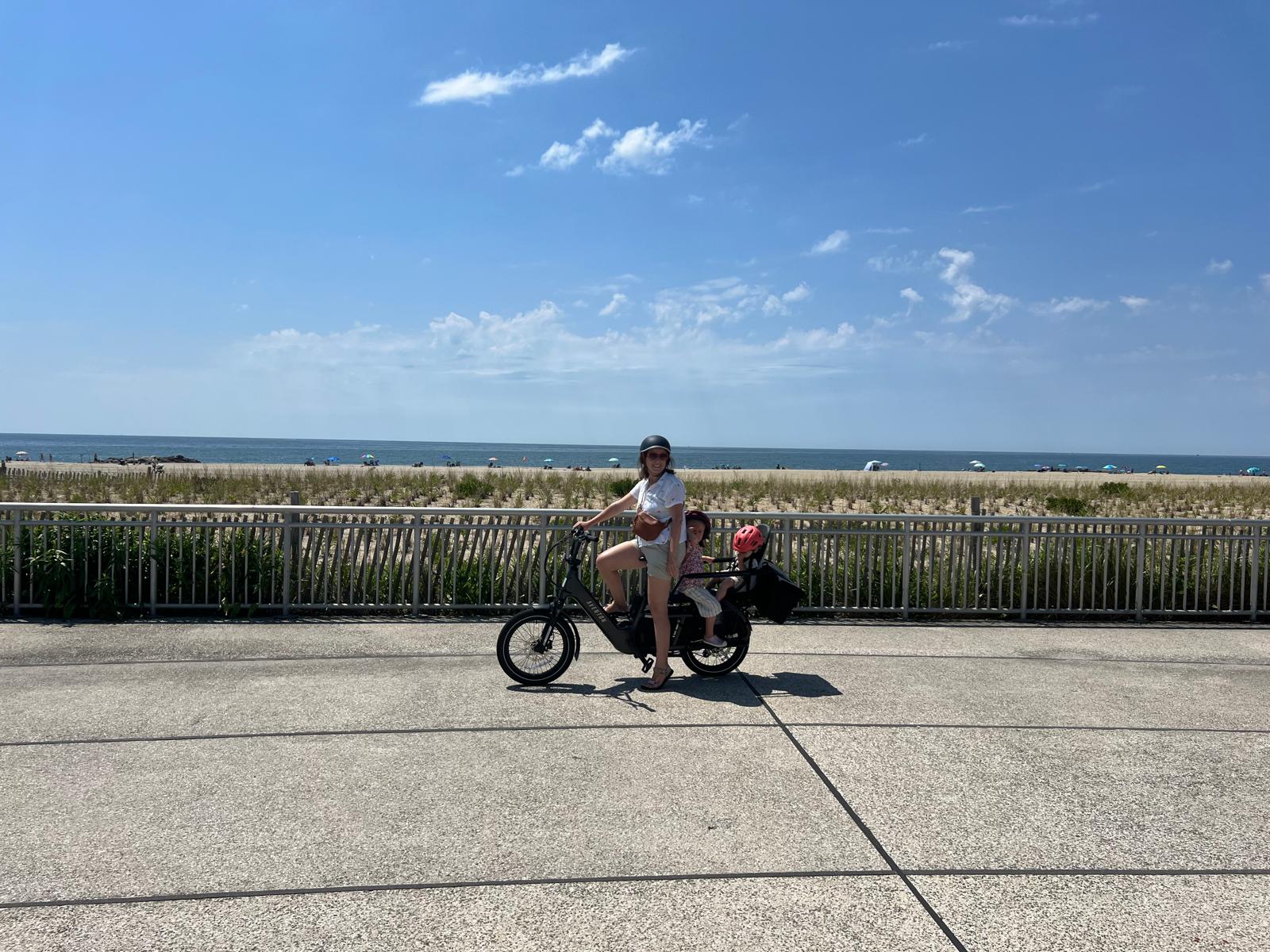 Giulia riding solo with both kids along the Rockaway boardwalk, ocean in the background